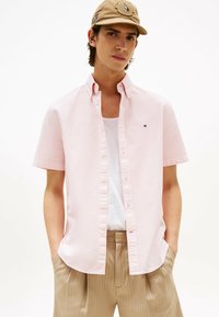 Jasnoróżowa koszula z krótkim rękawem, zapinana na guziki, z klasycznym kołnierzykiem i małym detalem logo. Połączona z beżowymi spodniami w paski.