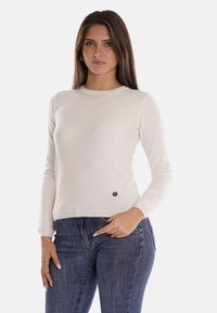Maglione beige chiaro con maniche lunghe, scollatura rotonda, polsini a costine e un piccolo logo sul lato sinistro. Abbinato a jeans blu scuro.