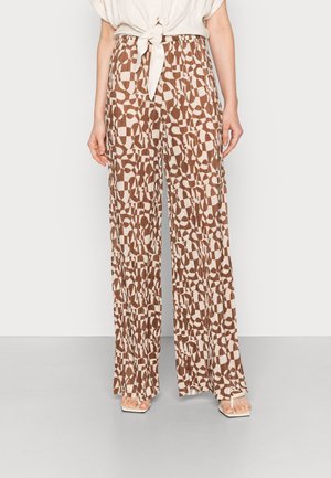Mujer con pantalones de pierna ancha en tonos marrón y beige con un patrón abstracto y una blusa crema anudada, de pie sobre un suelo gris claro.