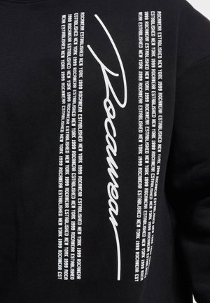 Sort sweatshirt med fed hvid tekst, herunder "Rocawear" i en stiliseret skrifttype og gentagne sætninger, der angiver "Etableret New York 1999."