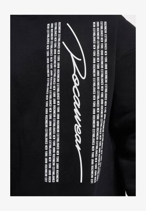 Fekete pulóver merész, fehér szöveggel, amely tartalmazza a "Rocawear" stilizált írásmódban, valamint az "Established New York 1999" ismétlődő kifejezéseit.