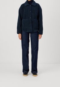 Granatowa polarowa kurtka z teksturowanym wykończeniem, z kołnierzykiem i kieszeniami z przodu, w połączeniu z ciemnymi dżinsami i czarnymi butami.