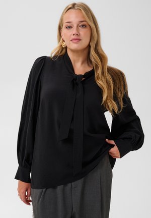 Femme aux longs cheveux blonds porte une blouse noire avec un nœud au col et des manches bouffantes, associée à un pantalon gris sur un fond uni.