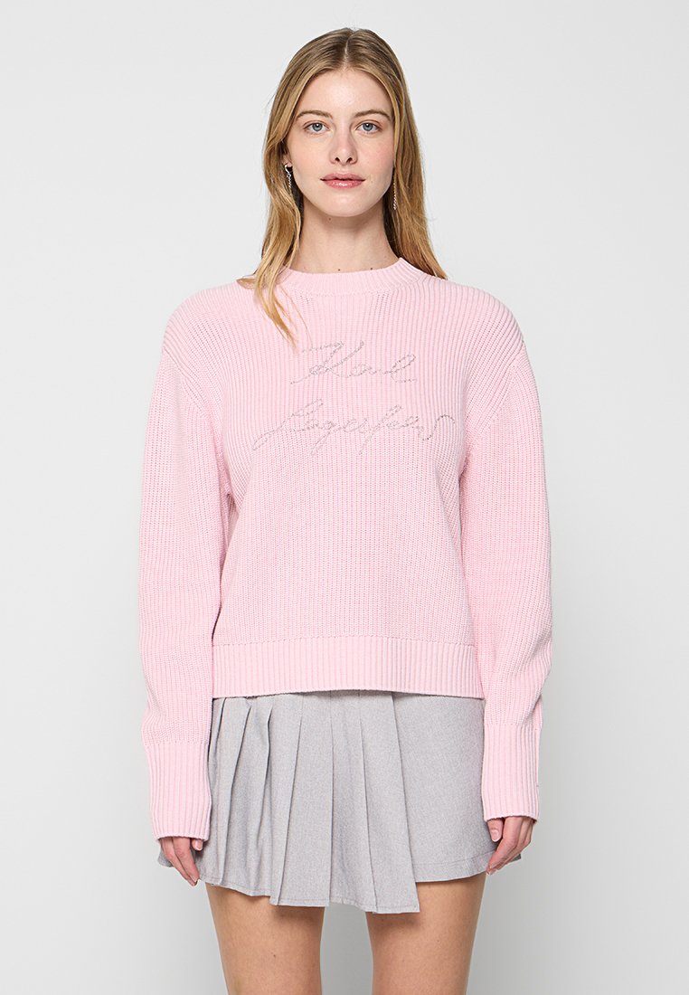 Pull en maille rose pâle avec texture côtelée et encolure ras du cou, arborant "Stay Together" brodé en argent. Porté par-dessus une jupe plissée grise.