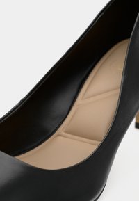 Décolleté in pelle nera con finitura liscia, caratterizzato da una soletta beige con un motivo geometrico e un leggero accento metallico sul tacco.