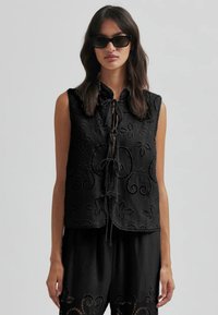 Top negru fără mâneci, cu un guler înalt, având broderie florală complicată, margine zimțată și un sistem de închidere cu legătură în față.