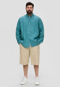 Turquoise linnen overhemd met knopen en lange mouwen, gecombineerd met beige shorts tot de knie. Het model draagt witte sneakers en heeft de handen in de zakken.
