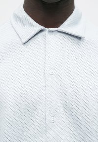 Lys grå polotrøye laget av strukturert stoff med mønster i netting, med spiss krage og tre knappelukking.