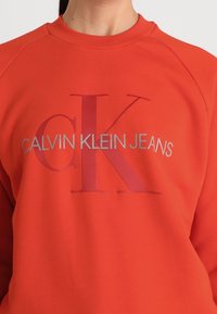 Sudadera roja con cuello redondo y mangas raglán. El logo exhibido dice "CALVIN KLEIN JEANS" con una gran 'K' en color contrastante.