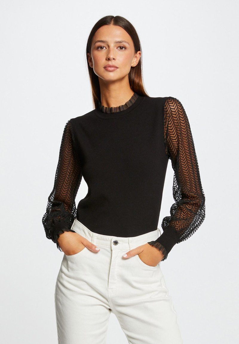 Top nero a maniche lunghe con maniche trasparenti a motivo herringbone testurizzato e dettagli di rouches sul collo e sui polsini. Abbinato a pantaloni bianchi.