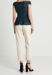 Haut péplum en dentelle turquoise à manches épaules dénudées, associé à un pantalon en velours côtelé crème et des sandales à talons noires. Vue de dos de la tenue.