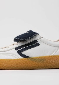 Hvid sneaker med en marineblå stofflap, tekstureret gummisål, bølgede marineblå accenter på siden og beige snørebånd.