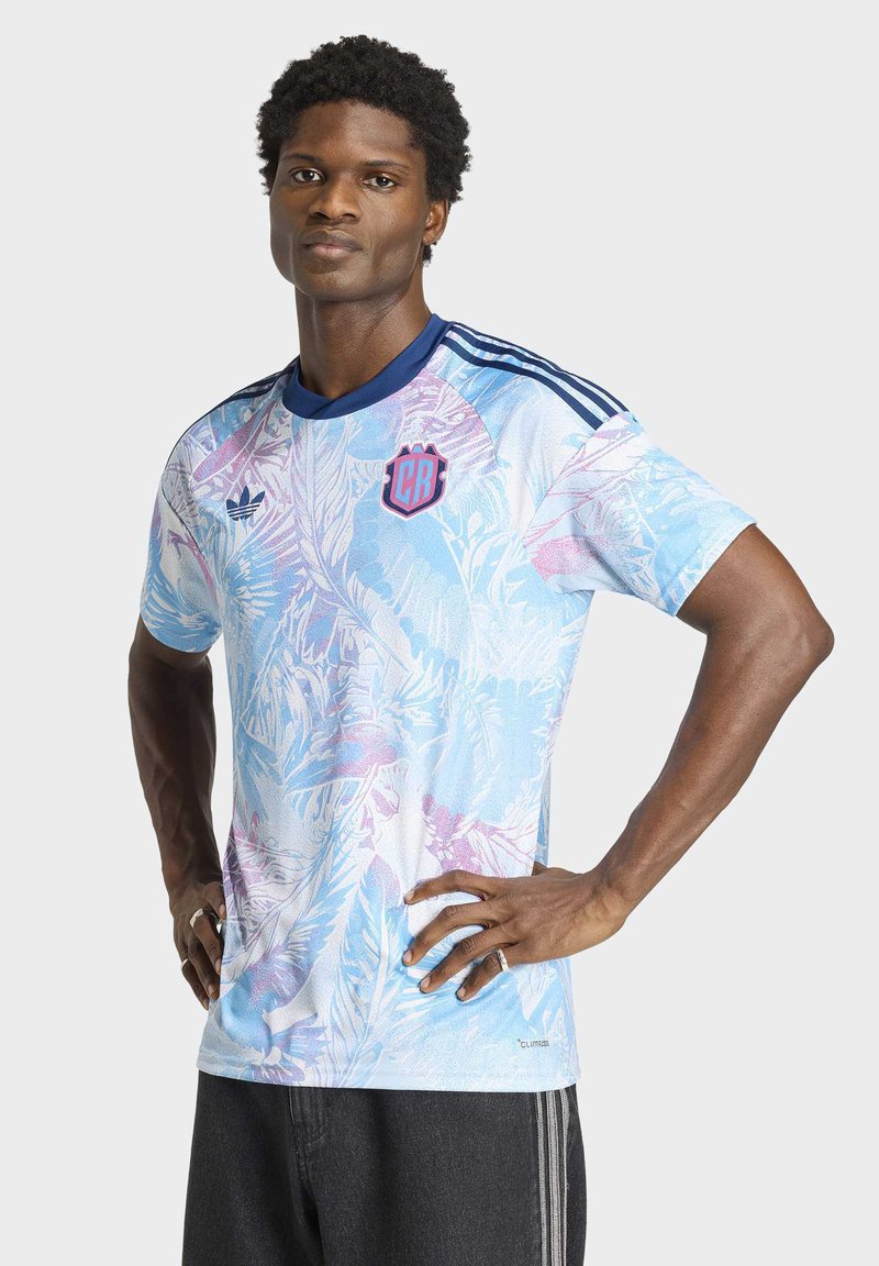 Junger Mann mit in die Hüften gestützten Händen, der ein kurzärmliges Sporttrikot in Pastellblau und Rosa mit dunklen Streifen auf den Schultern trägt.