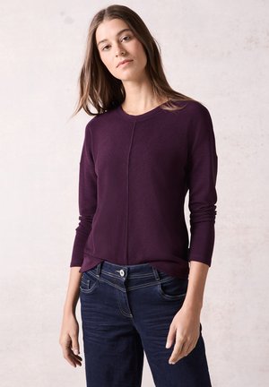 Chemise à manches longues texturée violette avec un col rond et des coutures détaillées, accompagnée de jeans bleu foncé avec des surpiqûres contrastantes.