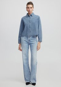 Camicia in denim blu con bottoni sul davanti, maniche lunghe e dettagli ricamati; abbinata a jeans in denim chiaro a zampa.