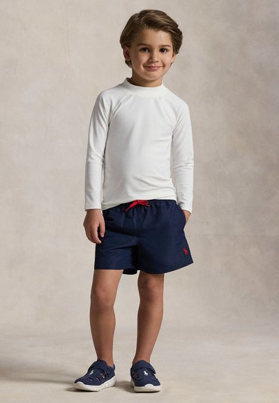 Junger Junge steht, trägt ein weißes Langarmshirt, marineblaue Shorts mit rotem Kordelzug und marineblaue Sneakers mit weißen Sohlen.