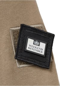 Beige stof met een zachte textuur heeft een zwart label met "WEEKEND OFFENDER" in het wit, omrand door een geweven zwart kader.