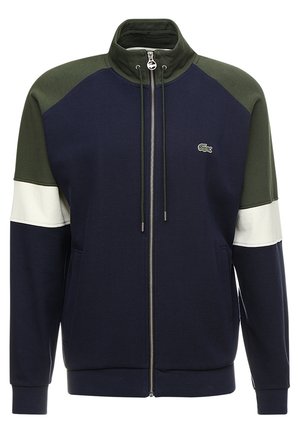 Veste zippée bleu marine, vert olive et blanche avec col haut, poches avant, cordons de serrage et petit logo crocodile vert sur la poitrine.