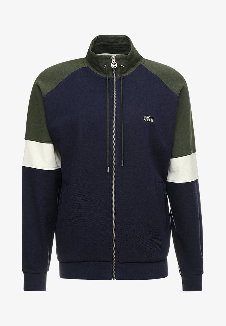 Veste zippée bleu marine, vert olive et blanche avec col haut, poches avant, cordons de serrage et petit logo crocodile vert sur la poitrine.