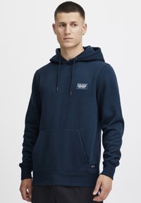 Felpa in cotone blu navy con tasca frontale, cappuccio regolabile con lacci e patch logo sul petto. Dotata di polsini e orlo a coste.