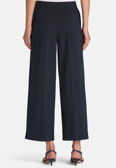 Nő sötétkék, széles szárú, rövidített nadrágban és fekete, magassarkú szandálban, hátulnézetben, fehér háttér előtt.