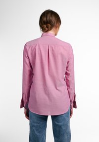 Žena s hnědými vlasy v drdolu, oblečená v růžové dlouhé košili a modrých džínách, stojící zády k bílé stěně.