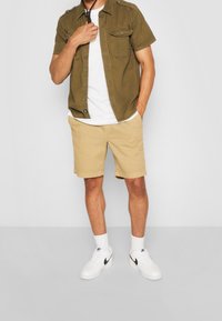 Homme portant une chemise à manches courtes olive à boutonnage, un t-shirt blanc, un short beige, des chaussettes blanches et des baskets blanches Nike, se tenant avec une main dans la poche.