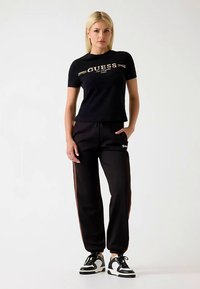 T-shirt en coton noir avec le logo « GUESS » en doré, associé à un pantalon de survêtement noir avec une bande latérale et un logo, complété par des baskets noires.