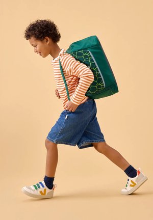 Grüner Rucksack mit geometrischem Muster, orange-weiß gestreiftes Langarmshirt, Jeansshorts und bunte Turnschuhe. Kind geht.
