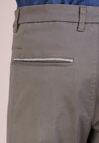 Šedé látkové kalhoty s kapsou na zadní straně se pruhovaným otvorem, hladkou texturou a jemným prošíváním pro přidaný vizuální zájem.