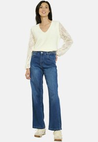 Suéter blanco de cuello en v con mangas de encaje, combinado con jeans acampanados azules. La modelo lleva zapatillas de color claro. Diseño sencillo y ajuste cómodo.