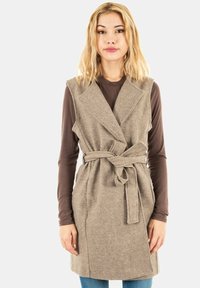 Beżowa kamizelka z wełny z wplecionymi włóknami, z fasonem kopertowym, głębokim dekoltem w kształcie litery V i paskiem w talii. Posiada boczne kieszenie i strukturalne wykończenie.