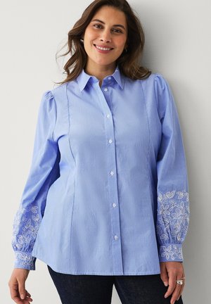 Kvinde smilende iført en lyseblå skjorte med knapper og hvid blomsterbroderi på puffede lange ærmer samt mørke bukser.