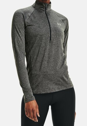 Kobieta ubrana w dopasowany szary sportowy top z długim rękawem i suwakiem do ćwiartki oraz czarne legginsy, stoi na jednolitym tle.