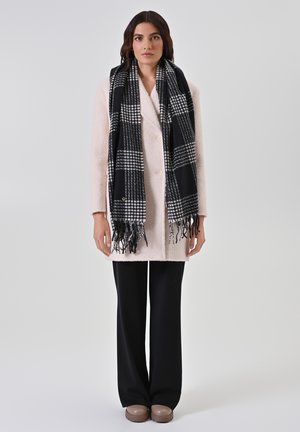 Lichtroze, dubbelzijdige jas van gestructureerde stof, gecombineerd met een zwart-wit geblokt sjaal met franjes. Zwarte broek en schoenen.
