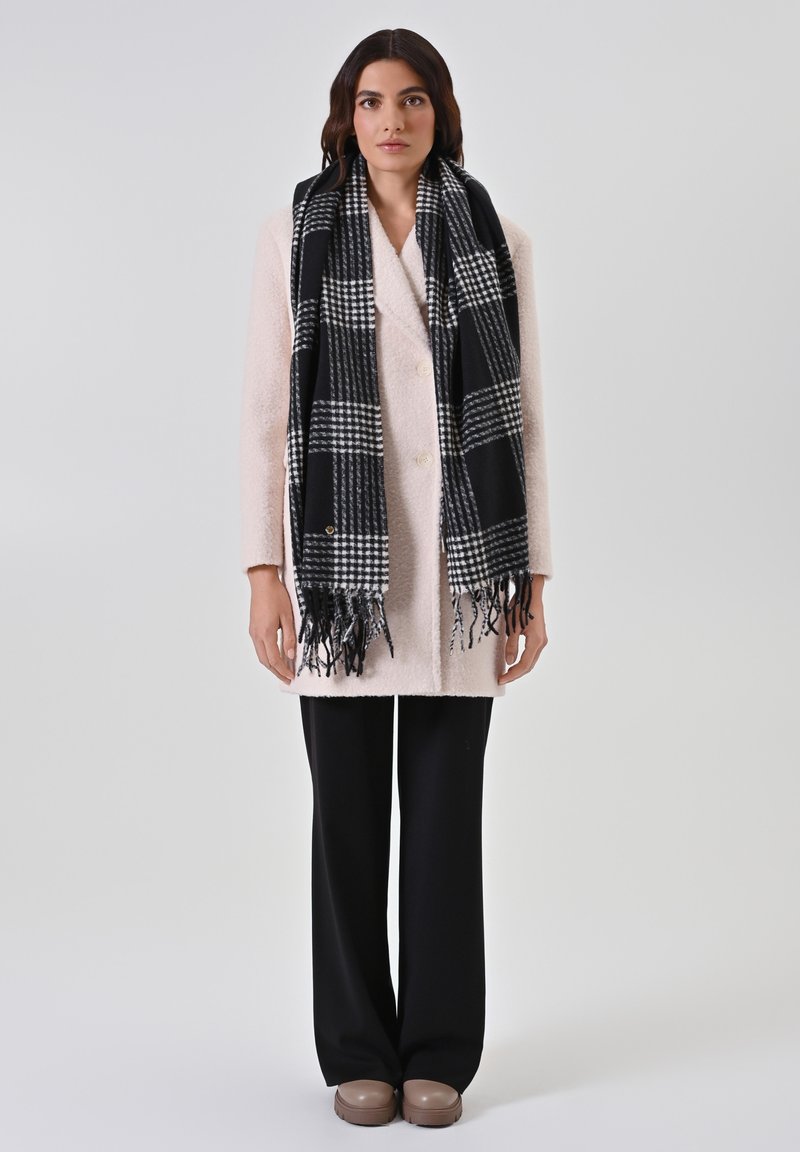 Lichtroze, dubbelzijdige jas van gestructureerde stof, gecombineerd met een zwart-wit geblokt sjaal met franjes. Zwarte broek en schoenen.