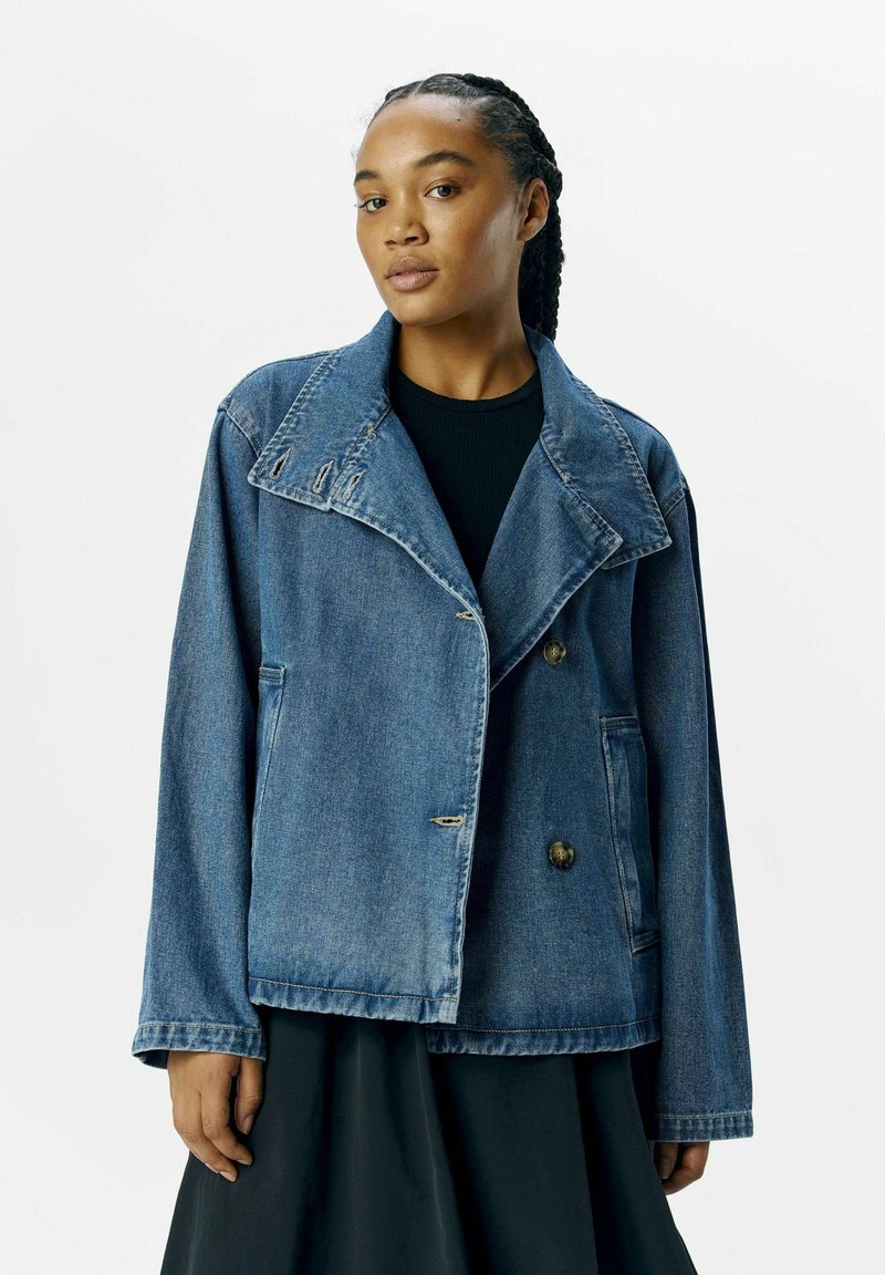 Veste en denim avec un col cranté, des boutons double boutonnage, deux poches latérales et une coupe légèrement oversize en délavage bleu moyen.