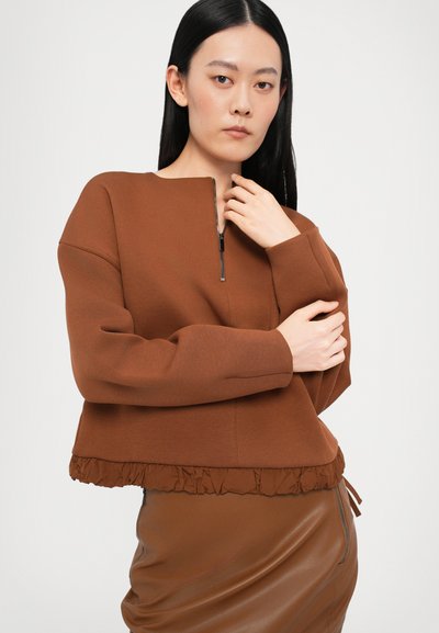 Rjav skrajšan pulover z visokim ovratnikom, ki ima sprednji zadrgo in teksturiran volanček na robu. Gladka blaga in sproščen kroj.