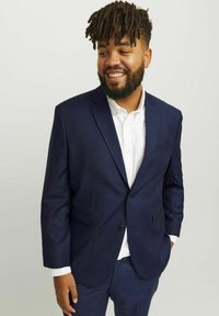 Navy blue suit with a tailored fit, single-button closure, and two front pockets. Worn over a white collared shirt, smooth fabric texture.