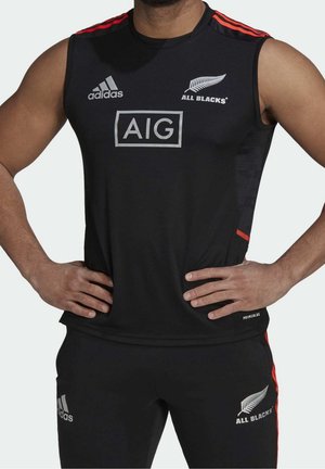 Męski model noszący czarną sportową koszulkę bez rękawów i spodnie z logo Adidas, srebrnymi paprociowymi emblemami All Blacks oraz czerwonymi paskami na ramionach i bokach.