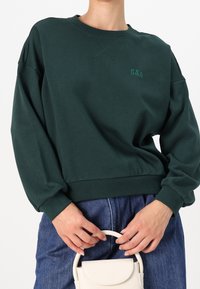 Sweat-shirt vert foncé avec ourlet et poignets côtelés, arborant un petit logo "GAP" vert. Porté avec un jean taille haute et un petit sac à main blanc.