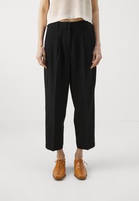 Schwarze maßgeschneiderte Hose mit einem entspannten Schnitt, verziert mit Falten und einer glatten Textur, kombiniert mit hellbraunen Schnürschuhen.