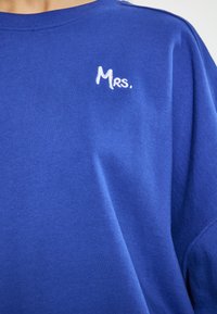 Királykék pamut póló, fehér hímzett "Mrs." felirattal. A dizájn környakú és laza fazonú.