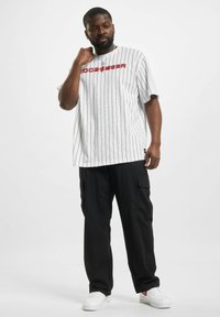 T-shirt blanche à rayures avec logo "Rocawear" rouge, associé à un pantalon cargo noir et des baskets blanches. Manches courtes et coupe décontractée.