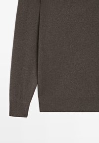 Manica e parte inferiore di un maglione a maglia marrone visibili su uno sfondo bianco, che mostrano i polsini a coste e la texture del tessuto.