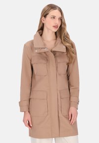 Knælang beige frakke med høj krave, dobbelte brystlommer og trykknaps-/knapdetaljer. Fremstillet af glat stof med en afslappet pasform.