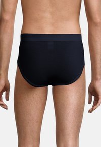 Man iförd svarta kalsonger, stående med ryggen mot kameran, armarna avslappnat vid sidorna, mot en enfärgad bakgrund.