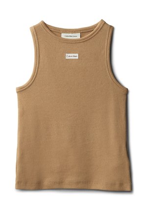 Jong meisje staand, gekleed in een bruin mouwloos topje, lichtblauwe wijde jeans en zwarte sandalen, tegen een effen grijze achtergrond.