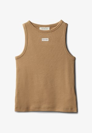 Jong meisje staand, gekleed in een bruin mouwloos topje, lichtblauwe wijde jeans en zwarte sandalen, tegen een effen grijze achtergrond.