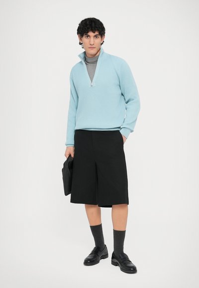 Pull bleu clair à fermeture éclair avec texture côtelée, associé à un short noir jusqu'aux genoux. Des mocassins noirs et des chaussettes grises complètent le look.